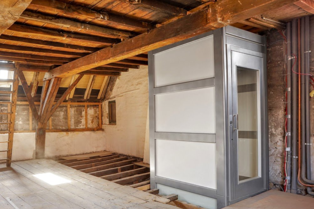 Ein moderner, grauer Aufzug mit Glastür, der in einem rustikalen Dachboden mit sichtbaren Holzbalken installiert ist. Das Bild zeigt die praktische Integration von einem Aufzug für den Dachboden bei der Altbausanierung.